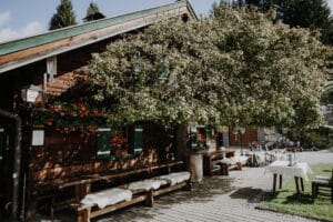 Eine rustikale Holzhütte mit grünen Fensterläden liegt unter einem großen Baum und bietet die perfekte Kulisse für eine Hochzeit. Rote Blumen schmücken die Fenster, während lange Bänke mit Fellüberwürfen und ein Tisch mit weißer Tischdecke auf der Terrasse auf die Gäste warten. Es ist ein sonniger Tag in dieser idyllischen Umgebung.