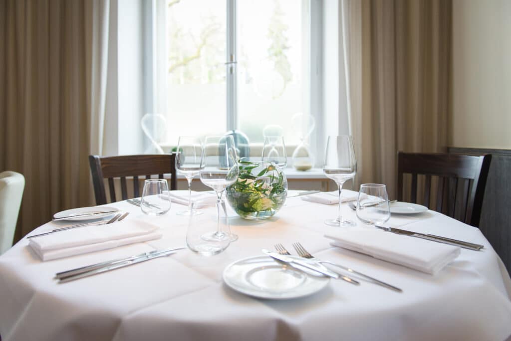 Ein runder Esstisch ist elegant mit einer weißen Tischdecke, Servietten, Besteck und leeren Weingläsern gedeckt. Eine kleine Glasschale mit Grünpflanzen ist perfekt für Hochzeitsfeierlichkeiten und dient als Tischdekoration. Der Tisch steht in der Nähe eines Fensters mit hellen Vorhängen und schafft so ein ideales Ambiente für Hochzeiten.