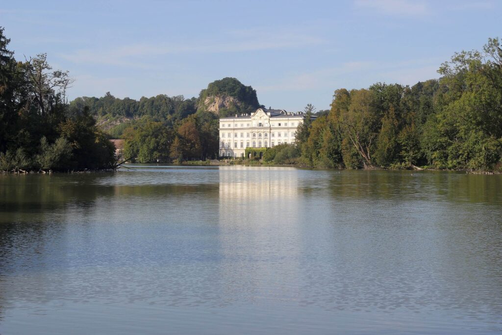 Ein großes weißes Herrenhaus liegt hinter einem ruhigen See und bietet eine bezaubernde Kulisse für eine Traumhochzeit. Umgeben von üppigen grünen Bäumen und Hügeln unter einem klaren blauen Himmel schimmert die Spiegelung des Gebäudes auf dem ruhigen Wasser und schafft eine perfekte Kulisse für diejenigen, die heiraten möchten.
