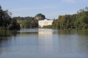 Ein großes weißes Herrenhaus liegt hinter einem ruhigen See und bietet eine bezaubernde Kulisse für eine Traumhochzeit. Umgeben von üppigen grünen Bäumen und Hügeln unter einem klaren blauen Himmel schimmert die Spiegelung des Gebäudes auf dem ruhigen Wasser und schafft eine perfekte Kulisse für diejenigen, die heiraten möchten.