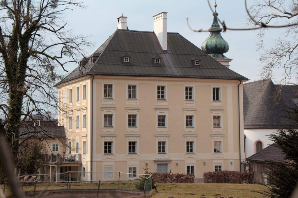 Ein großes, hellgelbes Gebäude mit zahlreichen Fenstern steht neben einer kleinen Kapelle mit Kuppeldach, ideal für eine intime Hochzeit. Kahle Bäume und ein eingezäunter Hof im Vordergrund verstärken die ruhige Atmosphäre unter dem bedeckten Himmel – eine perfekte Kulisse für eine durchdachte Hochzeitsplanung.