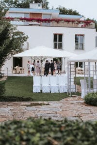 Eine Gruppe von Menschen versammelt sich unter weißen Sonnenschirmen auf einer Wiese vor einem modernen weißen Gebäude mit blumengeschmückten Balkonen. Im Vordergrund sind leere weiße Stühle aufgestellt, die auf Hochzeitsvorbereitungen hindeuten und eine elegante Hochzeitsveranstaltung suggerieren.