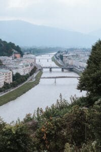 Blick auf eine Stadt, durch die ein Fluss fließt, perfekt für alle, die von einer Hochzeit am Wasser träumen. Mehrere Brücken überspannen den Fluss, gesäumt von charmanten Gebäuden und viel Grün. Im Hintergrund bilden sanfte Hügel eine bezaubernde Kulisse unter einem bewölkten Himmel.