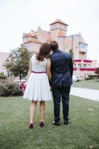 Ein Paar steht auf einer Wiese, die Hände ineinander verschränkt, als sie sich auf den Weg zur Hochzeit machen. Die Frau in einem fließenden weißen Kleid und der Mann in einem eleganten dunklen Anzug blicken auf Gebäude und Bäume, und ein Hauch von Romantik fängt diesen kostbaren Moment ein, bevor die Feierlichkeiten beginnen.