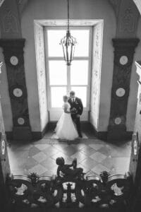 Ein Schwarzweißfoto von Braut und Bräutigam an ihrem Hochzeitstag, die vor einem verzierten Fenster in einem historischen Gebäude stehen. Dekorative Wände unterstreichen die zeitlose Kulisse, während darüber ein Kronleuchter schimmert. Eine Statue ziert den Vordergrund auf einer großen Treppe.