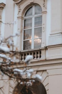 Ein klassisches Bogenfenster mit dekorativer Verzierung an einem hellen Gebäude lässt auf eine elegante Hochzeit schließen. Durch das Glas ist ein Kronleuchter sichtbar, der ein romantisches Licht verbreitet, während schneebedeckte Zweige den Blick im Vordergrund teilweise verdecken.