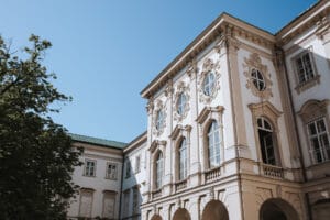 Ein historisches weißes Gebäude mit kunstvollen architektonischen Details, perfekt für eine Hochzeit, mit gewölbten Fenstern und Zierleisten, vor einem klaren blauen Himmel. Auf der linken Seite ist teilweise ein grüner Baum zu sehen, der den Charme noch verstärkt, ideal für die Hochzeitsplanung.