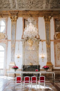 Ein üppig dekorierter Raum, der sich perfekt für die Hochzeitsplanung eignet, mit verzierten goldenen und weißen Wänden, einem großen Kronleuchter und einem kunstvollen Kamin. Vier elegante Stühle mit roten Kissen umgeben einen langen weißen Tisch, der mit rosa Blumen geschmückt ist und den idealen Rahmen für jede Hochzeitsfeier bildet.