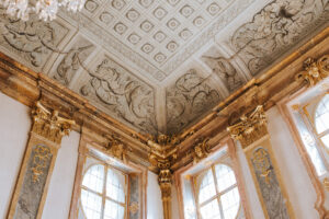 Das kunstvolle Innere dieses historischen Gebäudes, ideal für eine Hochzeit, verfügt über eine detailliert bemalte Decke mit komplizierten Mustern, große Bogenfenster und vergoldete Säulen. Natürliches Licht erhellt den Raum und hebt seine eleganten architektonischen Merkmale hervor, die perfekt zum Heiraten sind.