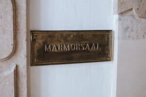 Auf einer Messingtafel an einer hellen Wand steht „Marmorsaal“ mit dezenten Marmordetails – ein perfekter Ort zum Heiraten, der Eleganz ausstrahlt und ideal für jede Hochzeitsplanung ist.