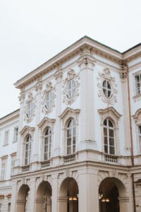 Ein historisches weißes Gebäude mit kunstvollen architektonischen Details, perfekt für alle, die eine Hochzeitsfeier planen. Es verfügt über runde und gewölbte Fenster, dekorative Zierleisten und eine Reihe gewölbter Eingänge im Erdgeschoss. Der bewölkte Himmel verleiht dieser eleganten Umgebung eine gedämpfte Atmosphäre.