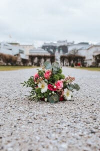 Ein Strauß rosa Rosen, weißer Gänseblümchen und Grünpflanzen liegt auf einem Kiesweg, vor der verschwommenen Kulisse eines Parkgeländes und von Gebäuden unter einem bewölkten Himmel – eine elegante Note für jede Hochzeitskulisse.