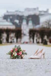 Ein Paar beige High Heels steht neben einem farbenfrohen Strauß rosa und weißer Blumen auf einem Kiesweg und schafft vor der verschwommenen Kulisse eines historischen Gebäudes und üppigen Grüns die perfekte Szene für eine Hochzeit.