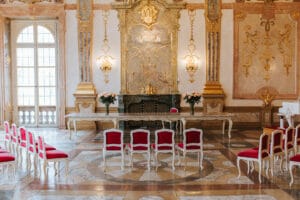 Der elegante Raum, der sich ideal für eine Hochzeit eignet, verfügt über einen langen, mit Blumenarrangements geschmückten Tisch, umgeben von weißen Stühlen mit roten Kissen. Mit kunstvollen goldenen Akzenten, großen Fenstern und aufwendigen Wanddesigns schafft er eine luxuriöse Atmosphäre, die perfekt für jede Hochzeitsfeier ist.