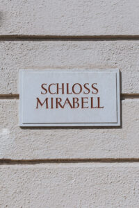Eine weiße Steintafel mit roter Schrift trägt die Aufschrift „Schloss Mirabell“ und ist an einer strukturierten, hellen Wand angebracht. Sie bietet den perfekten Hintergrund für Ihre Hochzeitsplanung.