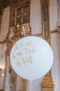 Ein weißer Ballon mit der goldenen Aufschrift „Du & Ich bis ans Ende der Welt“ schwebt anmutig in einem reich verzierten Raum, der sich ideal für eine Hochzeitsplanung eignet und mit hohen Fenstern, Kronleuchtern und aufwendigen Zierleisten geschmückt ist.