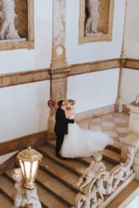 Braut und Bräutigam küssen sich auf einer verzierten Treppe und verkörpern damit den perfekten Hochzeitsmoment. Das weiße Kleid der Braut bildet einen Kontrast zum dunklen Anzug des Bräutigams, umgeben von klassischer Architektur, geschmückt mit Skulpturen und detailreichem Mauerwerk. Ein Blumenstrauß hängt elegant neben ihnen.