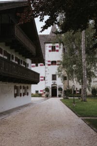 Ein charmantes Gebäude mit weißen Wänden und roten Fensterläden, perfekt für eine Hochzeit, mit einem turmähnlichen Teil. Ein Kiesweg führt zum gewölbten Eingang und die Umgebung umfasst einen Rasen, hohe Bäume und einen Laternenpfahl. Ideal für diejenigen, die ihre Traumhochzeit planen.