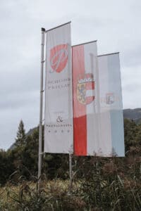Drei vertikale Flaggen wehen auf einer Rasenfläche. Auf einer steht: „Schloss Prielau Hotel & Restaurants“ mit Wappen. Eine andere Flagge zeigt rot-weiße Streifen mit Wappen, während die dritte ein „H“ auf blau-weißem Hintergrund zeigt, was auf den exquisiten Charme des Veranstaltungsortes für Hochzeitsplanung hinweist.