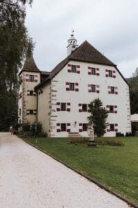 Ein großes, historisches weißes Gebäude mit roten Fensterläden und einem spitzen Turm steht inmitten eines grünen Rasens, perfekt für eine Traumhochzeit. Ein Kiesweg führt zum Eingang, flankiert von einer kleinen Statue und Bäumen unter einem bewölkten Himmel.