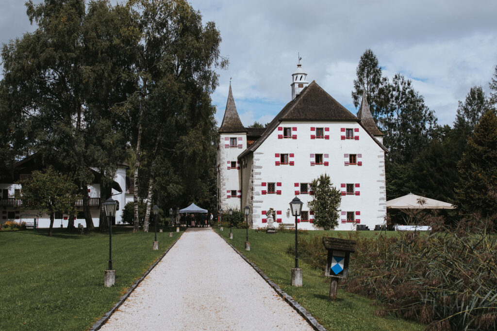 Ein malerischer Weg führt zu einem historischen weißen Schloss mit roten Fensterläden, perfekt für eine märchenhafte Hochzeit. Umgeben von üppigen grünen Rasenflächen und hohen Bäumen bietet es eine bezaubernde Kulisse für Ihre Hochzeitsplanung. Über Ihnen liegt ein teilweise bewölkter Himmel, und in der Nähe des Eingangs sind einige Menschen zu sehen.