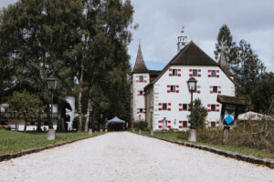Ein weißes Schloss mit rot umrandeten Fenstern, perfekt für eine Hochzeit, steht in einer üppigen grünen Landschaft. Ein Kiesweg führt zum Eingang, gesäumt von Laternenpfählen und Bäumen. In der Nähe des Eingangs ist unter einem bewölkten Himmel ein Baldachin zu sehen, der eine idyllische Kulisse für Ihre Hochzeitsplanung bietet.
