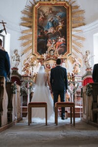 Braut und Bräutigam stehen Seite an Seite am Altar einer reich verzierten Kirche und sind bereit zu heiraten. Sie blicken auf ein großes, farbenfrohes Gemälde, das von goldenen Strahlen und Engelsstatuen umgeben ist. Im Vordergrund sind Holzbänke zu sehen, die die feierliche und heilige Atmosphäre der Hochzeit verstärken.