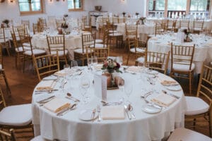 Ein wunderschön eingerichteter Bankettsaal, perfekt für eine Hochzeit, mit runden, weiß gedeckten Tischen, die mit ordentlich angeordnetem Besteck, Gläsern und Blumenschmuck geschmückt sind. Goldene Stühle umgeben die Tische, während natürliches Licht durch große Fenster hereinströmt.