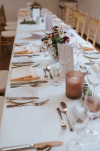 Ein langer, elegant gedeckter Esstisch mit weißer Tischdecke, gefalteten Servietten und arrangiertem Besteck ruft den Charme einer perfekten Hochzeitskulisse hervor. Es gibt Blumengestecke, Weingläser und dekorative Kerzen. Goldfarbene Stühle säumen die Seiten und verstärken die formelle und einladende Atmosphäre für Hochzeiten.