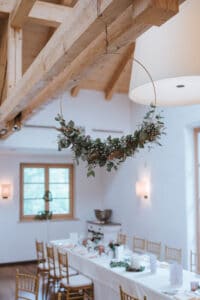 Ein rustikales Esszimmer mit Holzdecke und weißen Wänden ist perfekt für die Hochzeitsplanung. Es verfügt über einen langen, für ein Essen gedeckten Tisch, während ein runder Blumenkranz mit Grünpflanzen und weißen Blumen von oben herab hängt. Holzstühle umgeben den elegant dekorierten Tisch, ideal für eine intime Hochzeit.