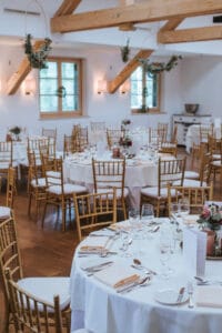 Ein wunderschön dekorierter Bankettsaal, der sich perfekt für eine Hochzeit eignet. Er bietet runde Tische mit weißen Tischdecken, elegantem Geschirr und Blumendekorationen. Der Raum verfügt über Holzbalken, sanfte Beleuchtung und von der Decke hängende Gründekorationen, ideal für Ihre Hochzeitsplanung.