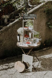 Ein kleiner Tisch im Freien ist elegant für eine Prosecco-Bar arrangiert, perfekt für eine Hochzeitsfeier. Das Schild „Pimp Your Prosecco“ lädt die Gäste ein, ihre Getränke mit Beeren, Kräutern und Blumen zu personalisieren. Eine kleine Tafel und ein Spiegel schmücken den Tisch, ergänzt durch rustikale Holzscheiben und Steintreppen im Hintergrund.