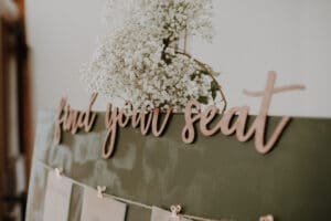 Ein Sitzplan mit grünem Hintergrund und der hölzernen Aufschrift „Finden Sie Ihren Sitzplatz“ ist perfekt für die Hochzeitsplanung. Ein Strauß zarter weißer Blumen ziert die Buchstaben, während darunter Papiere mit den Sitzordnungen der Gäste für eine elegante Hochzeit angebracht sind.