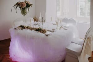 Ein runder Tisch, der exquisite Hochzeitsplanung verkörpert, ist für eine formelle Veranstaltung elegant mit weißem Tüll, Blumenarrangements und Kerzen geschmückt. Der Tisch ist mit Gläsern und Besteck gedeckt, umgeben von weiß bezogenen Stühlen mit Schleifen. Sanftes rosa Licht verleiht der Hochzeitskulisse eine romantische Note.