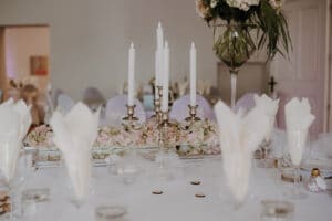 Ein wunderschön gedeckter Tisch mit einem silbernen Kerzenleuchter, der weiße Kerzen hält, umgeben von elegant gefalteten Servietten und Glaswaren, perfekt für die Hochzeitsplanung. Zarte rosa Blumen und eine hohe Vase mit Blumenarrangement verleihen dieser zauberhaften Hochzeitskulisse Raffinesse.