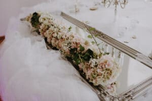 Ein dekoratives Tischmittelstück, perfekt für die Hochzeitsplanung, mit rosa und weißen Blumen, Grünpflanzen und Schleierkraut, alles arrangiert auf einem rechteckigen Spiegel mit verzierten Kanten. Der Tisch ist elegant mit weichem, weißem Tüllstoff drapiert.