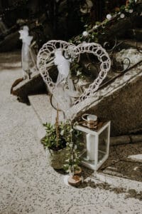 Ein dekoratives Herz aus gewebtem Material steht auf einer Steinoberfläche neben einer kleinen Topfpflanze und einer weißen Laterne mit einer Kerze darin, perfekt für eine intime Hochzeitsatmosphäre. Der Bereich ist mit weißem Stoff und Grün geschmückt und schafft eine charmante, rustikale Hochzeitsatmosphäre im Freien.