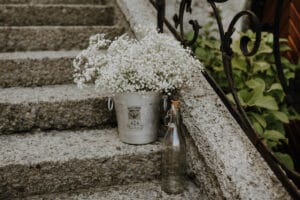 Ein silberner Eimer voller zarter weißer Blumen, perfekt für eine Hochzeit, steht auf Steinstufen neben einem dekorativen schwarzen Geländer. Eine klare Glasflasche mit Korken ergänzt die Szene und strahlt zeitlose Eleganz aus – die ideale Note für Ihre Hochzeitsplanung inmitten üppigen Grüns.