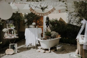 Ein Hochzeitsempfang im Freien besteht aus einem „Just Married“-Banner, einem Tisch mit Getränken, einer dekorierten Badewanne und einem kleinen Stand mit einem „Pimp Your Prosecco“-Schild. Die Szene ist mit Blumen und rustikalen Dekorationselementen geschmückt, perfekt für diejenigen, die schon weit in ihrer Hochzeitsplanung sind.