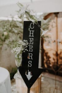 Ein schwarzes vertikales Holzpfeilschild mit „CHEERS“ in weißen Buchstaben weist den Weg zur Feier. Zwei Champagnergläser, gefüllt mit weißen Blumen und Schleierkraut, markieren einen Weg der Liebe. Der sanft verschwommene Hintergrund aus Grün und Holz deutet auf die zauberhafte Reise der Hochzeitsplanung hin.