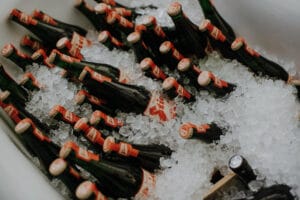 Mehrere dunkle Bierflaschen mit roten Etiketten und Verschlüssen liegen in einem großen Behälter, möglicherweise einer Badewanne, in zerstoßenem Eis. Die Szene lässt auf die Vorbereitungen für eine Hochzeitsfeier schließen, wobei das Eis die Getränke kühl und erfrischend hält – ideal für jede Hochzeitsfeier.