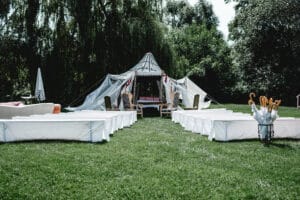 Eine skurrile Außenkulisse, perfekt für eine Hochzeit, mit einem Zelt inmitten der Natur. Weiße Bänke säumen einen grasbewachsenen Gang, der zum Zelt führt. Auf der rechten Seite sind Sonnenschirme aufgestellt und Bäume bilden eine üppige Kulisse für dieses ruhige Hochzeitsarrangement.