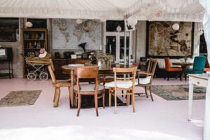 Ein Raum im Vintage-Stil mit eklektischen Möbeln, Holzstühlen und Tischen, perfekt für die Planung einer intimen Hochzeit. Karten und Kunstwerke schmücken die Wände, während im Hintergrund ein Kinderwagen zu sehen ist. Sanftes Licht verstärkt die gemütliche Atmosphäre.