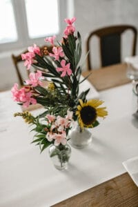 Ein kleines Blumenarrangement mit rosa Blumen, grünen Blättern und einer gelben Sonnenblume in einer Glasvase schmückt den Holztisch und passt perfekt zu einer Hochzeit. Es steht auf einem weißen Tischläufer in einem hellen Raum und verleiht Ihrem besonderen Tag Charme.