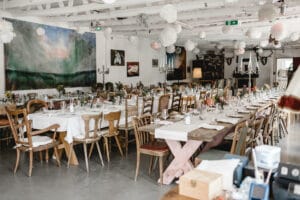 Ein geräumiger Innenbereich, der sich perfekt für eine Hochzeit eignet, mit langen Holztischen, die für ein Bankett gedeckt sind. Die Tische sind mit weißen Tischdecken, Kerzen und kleinen Blumenarrangements geschmückt. Kunstwerke schmücken die Wände und an der Decke sind freiliegende Balken sichtbar, die einen idealen Rahmen für die Hochzeitsplanung schaffen.