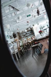 Ein rustikales Esszimmer mit einem langen Holztisch, der für eine Hochzeit gedeckt ist. Die Stühle passen nicht zusammen und die Decke ist mit dekorativen weißen Papierblumen geschmückt. Im Hintergrund sind Regale mit Glaswaren und Vasen zu sehen, die in ihrer Spiegelung den Charme der Hochzeitsplanung widerspiegeln.