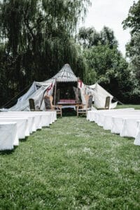 Ein großes Außenzelt auf einer Rasenfläche, perfekt für eine Hochzeit, mit weiß bedeckten Bänken, die zu einem offenen Eingang führen. Umgeben von hohen Bäumen schafft es eine ruhige, natürliche Atmosphäre, ideal für diejenigen, die heiraten möchten.