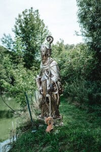 Eine goldene Statue einer verhüllten Gestalt, die einen Stab hält, steht auf Grasboden in der Nähe eines Teichs, umgeben von üppigen grünen Bäumen und Pflanzen. Ihre Präsenz verleiht der ruhigen Umgebung eine bezaubernde Note und macht sie zu einem perfekten Ort für romantische Hochzeitszeremonien. Am Sockel der Statue befindet sich eine kleine Holzskulptur.