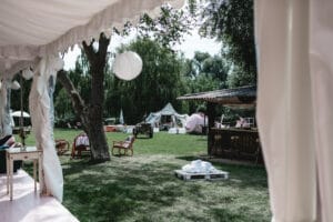 Eine skurrile Szene im Freien für eine Hochzeit, mit einer für die Veranstaltung geschmückten Rasenfläche. Papierlaternen baumeln von Bäumen inmitten eleganter Sitzgelegenheiten und charmanter Zelte. In der Nähe steht eine aus Holzpaletten gefertigte Bar, umgeben von Bäumen unter einem klaren Himmel.