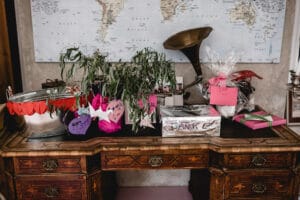 Ein Holzschreibtisch, geschmückt mit verschiedenen Geschenken, darunter eine eingewickelte Schachtel mit der Aufschrift „BANK“, eine Topfpflanze und kreativ verpackte Geschenke für eine Hochzeit. Im Hintergrund hängen ein Vintage-Grammophon und eine Weltkarte an der Wand und bilden eine elegante Bühne für diese besondere Feier.